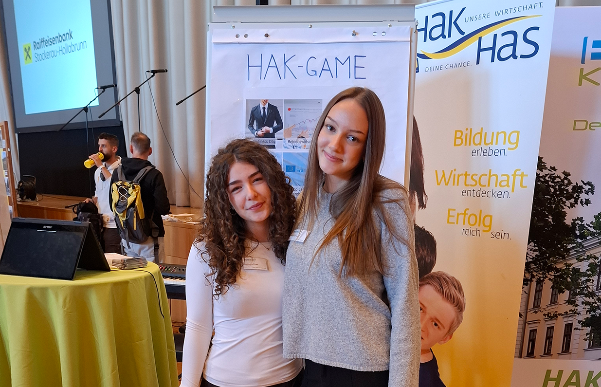Zwei junge Frauen vor HAK-GAME-Plakat und Werbebanner in Österreich